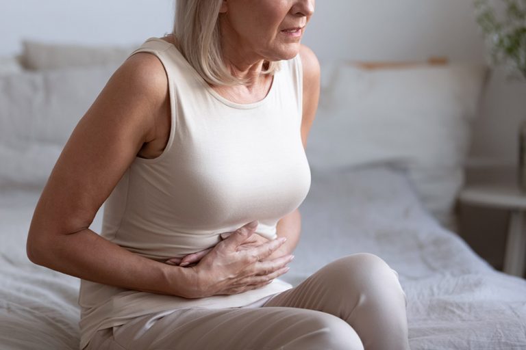 Unhealthy middle aged frowning woman sitting on bed, putting hands on belly, suffering from strong stomach ache. Unhappy older lady having painful feeling in abdomen, pancreatitis gastritis symptom.
