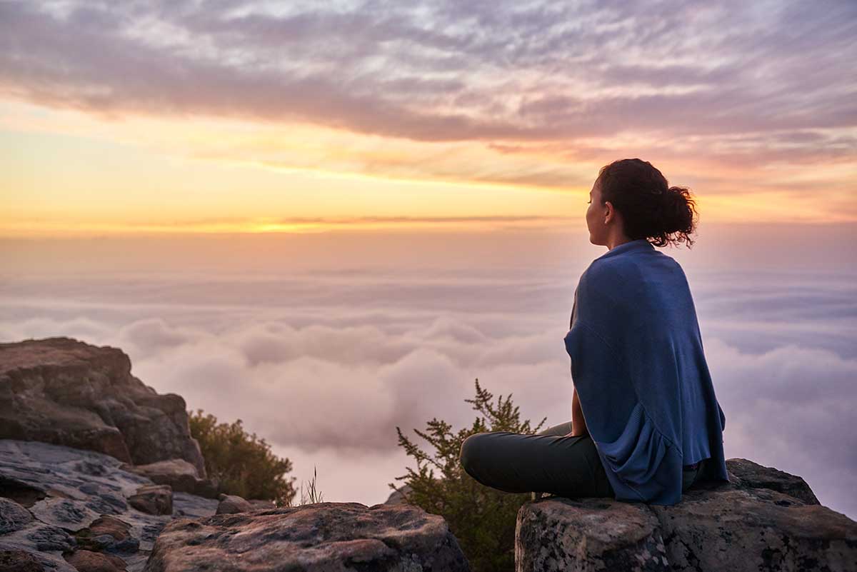 Rear view of a young woman sitting on a mountain top peacefully gazing at low-lying morning clouds and the pastel colours of a tranquil sunrise